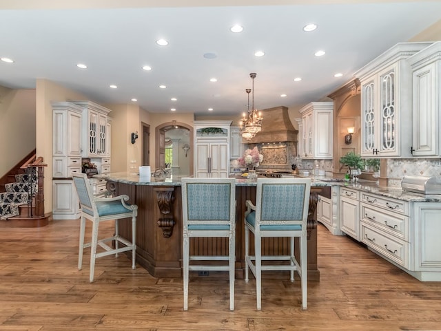 kitchen with premium range hood, glass insert cabinets, a large island with sink, and decorative light fixtures