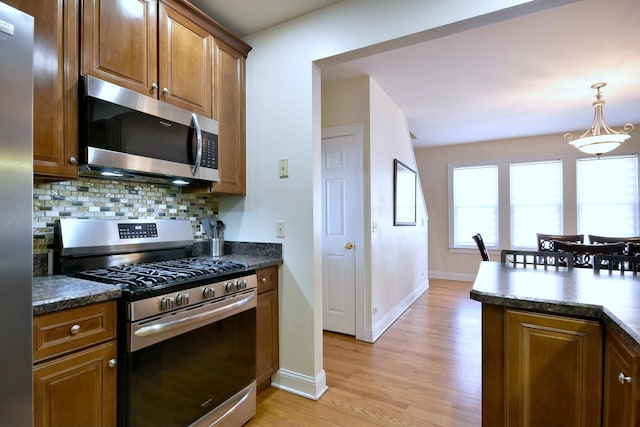 kitchen featuring light wood-style flooring, stainless steel appliances, hanging light fixtures, tasteful backsplash, and dark countertops