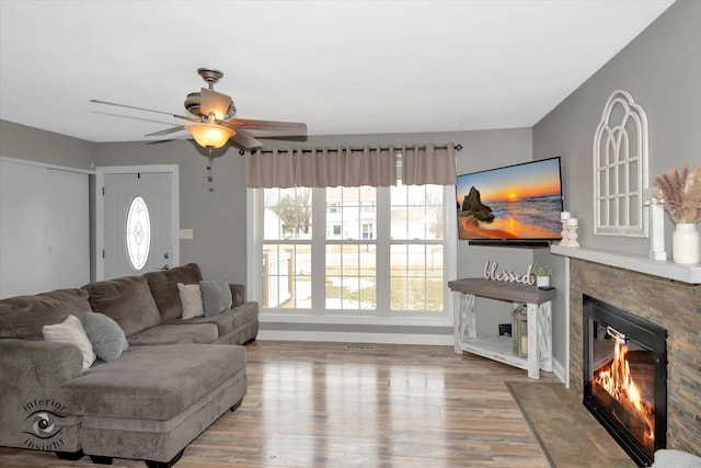 living room featuring wood finished floors, a glass covered fireplace, a ceiling fan, and baseboards