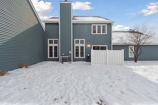 snow covered house with a chimney, cooling unit, and fence