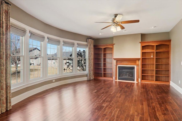 unfurnished living room featuring a ceiling fan, baseboards, dark wood-type flooring, and a glass covered fireplace