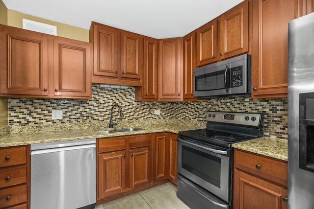 kitchen featuring brown cabinets, a sink, light stone counters, backsplash, and stainless steel appliances