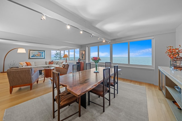 dining room featuring light wood-style flooring, a water view, baseboards, beamed ceiling, and rail lighting