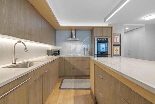 kitchen featuring modern cabinets, stainless steel double oven, light wood-type flooring, wall chimney range hood, and a sink