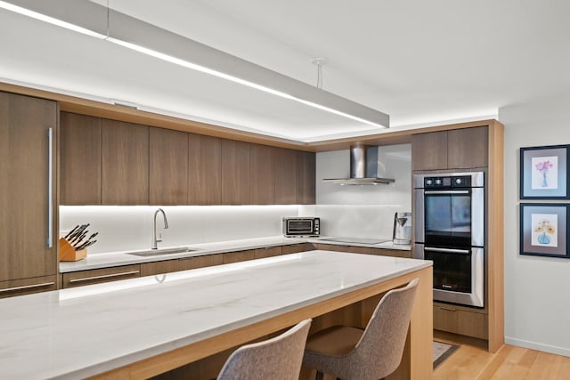 kitchen with stainless steel double oven, a breakfast bar area, black electric cooktop, a sink, and wall chimney range hood