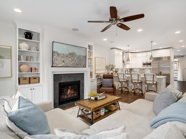 living area with a tiled fireplace, ceiling fan, ornamental molding, dark wood-type flooring, and recessed lighting