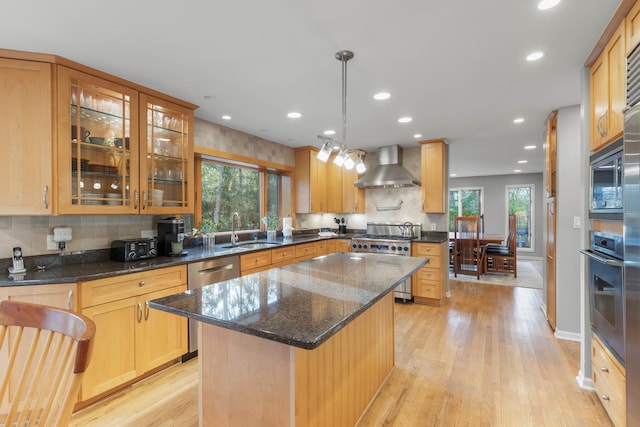 kitchen featuring wall chimney exhaust hood, appliances with stainless steel finishes, a center island, hanging light fixtures, and a sink