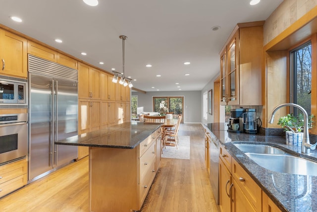 kitchen with dark stone counters, glass insert cabinets, built in appliances, hanging light fixtures, and a sink