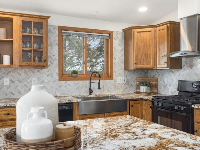 kitchen with light stone counters, glass insert cabinets, a sink, wall chimney range hood, and black appliances