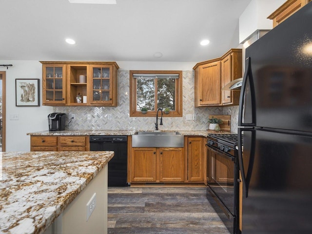 kitchen featuring glass insert cabinets, light stone counters, brown cabinets, black appliances, and a sink