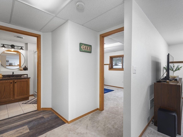 corridor with a paneled ceiling, baseboards, a sink, and light colored carpet