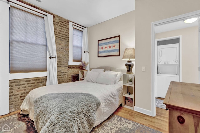 bedroom with brick wall, stacked washer and dryer, wood finished floors, and baseboards