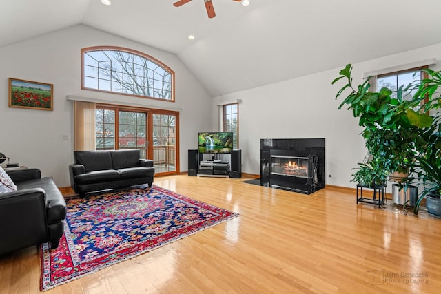 living room with high vaulted ceiling, wood finished floors, a ceiling fan, baseboards, and a glass covered fireplace