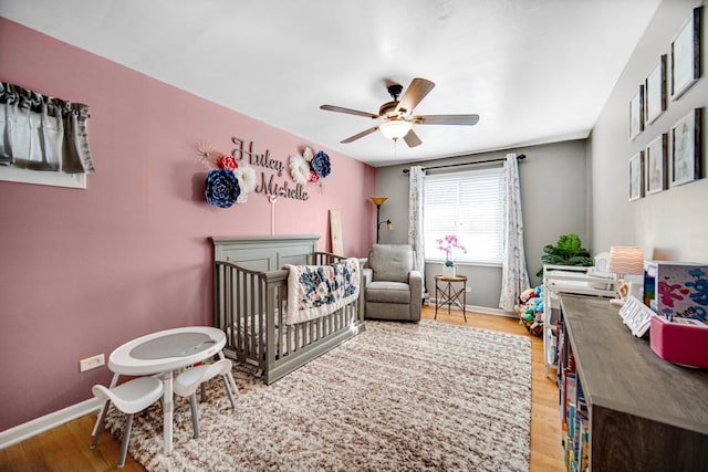 bedroom with a crib, ceiling fan, baseboards, and wood finished floors