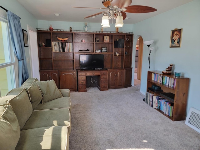 living area with arched walkways, ceiling fan, light colored carpet, visible vents, and built in study area