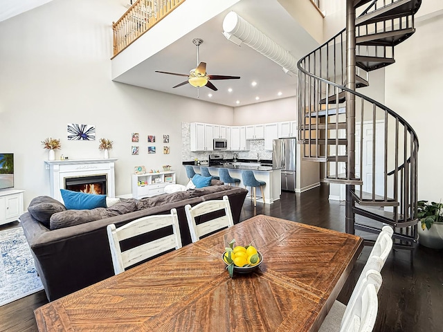 dining room with a warm lit fireplace, a high ceiling, dark wood-type flooring, a ceiling fan, and stairs