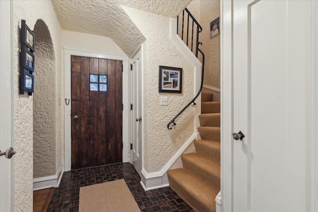 entryway with brick floor, a textured wall, baseboards, and stairs