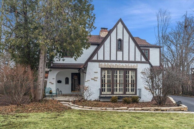 english style home featuring a chimney, stucco siding, a shingled roof, covered porch, and a front lawn