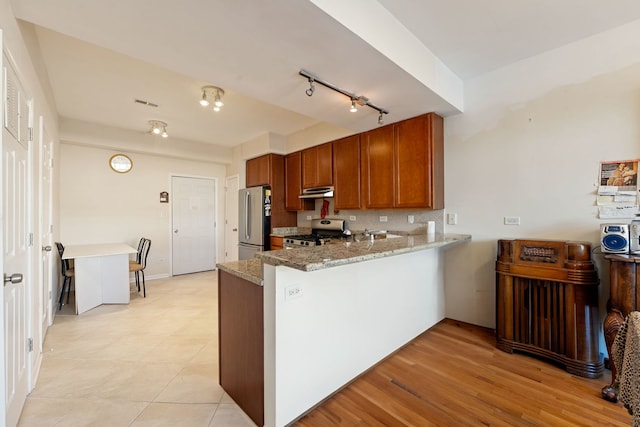 kitchen featuring decorative backsplash, appliances with stainless steel finishes, brown cabinetry, light stone countertops, and a peninsula