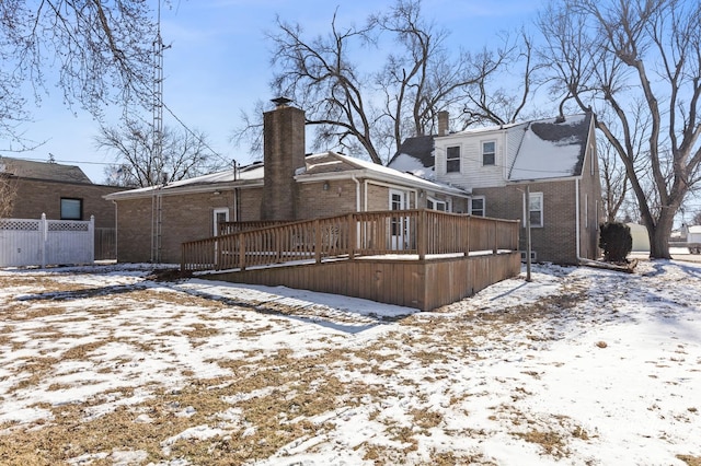 snow covered property featuring brick siding, a chimney, a wooden deck, and fence