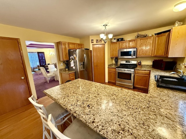 kitchen featuring brown cabinets, a peninsula, light stone countertops, stainless steel appliances, and a notable chandelier