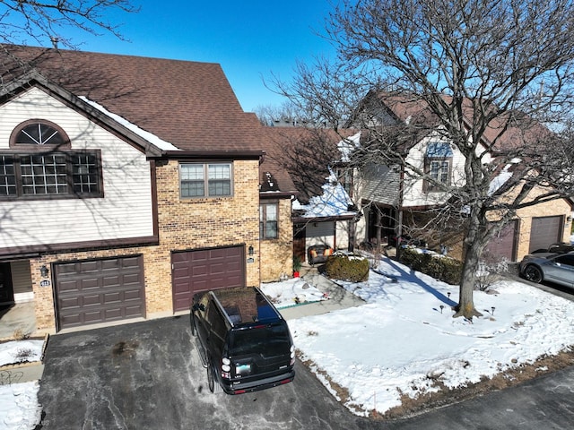 snow covered property featuring an attached garage, brick siding, a shingled roof, and aphalt driveway