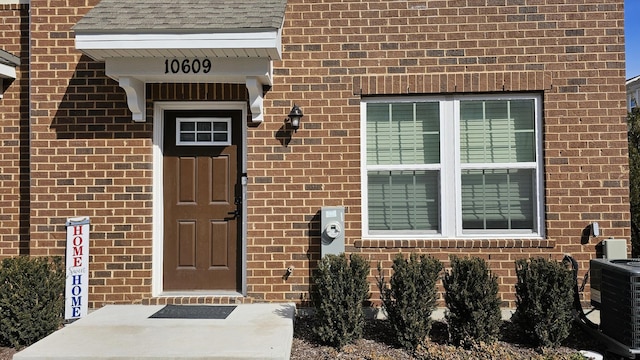 doorway to property featuring a shingled roof, brick siding, and central AC