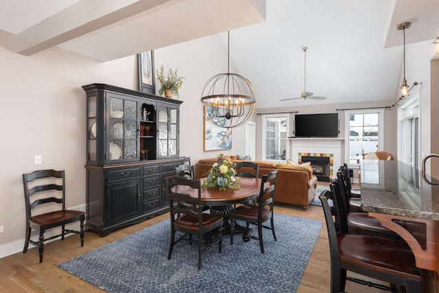 dining area with lofted ceiling with beams, light wood-style flooring, ceiling fan with notable chandelier, a fireplace, and baseboards