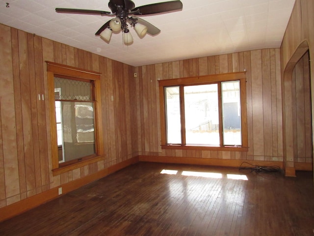 spare room featuring ceiling fan, arched walkways, wooden walls, baseboards, and dark wood-style floors