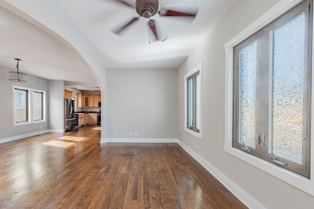 unfurnished living room featuring arched walkways, dark wood finished floors, a ceiling fan, and baseboards