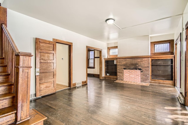 unfurnished living room featuring dark wood-style flooring, visible vents, baseboards, stairway, and a brick fireplace