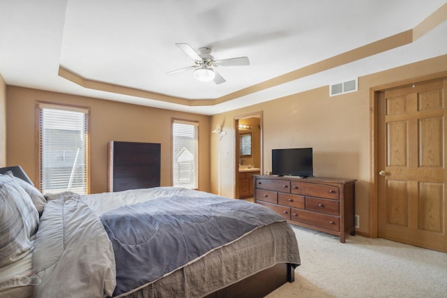 bedroom with light carpet, visible vents, a ceiling fan, a raised ceiling, and ensuite bath