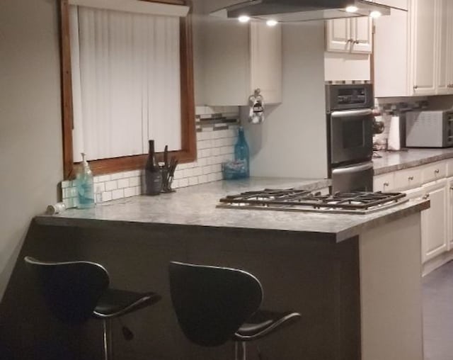 kitchen featuring tasteful backsplash, white cabinets, appliances with stainless steel finishes, a peninsula, and wall chimney range hood