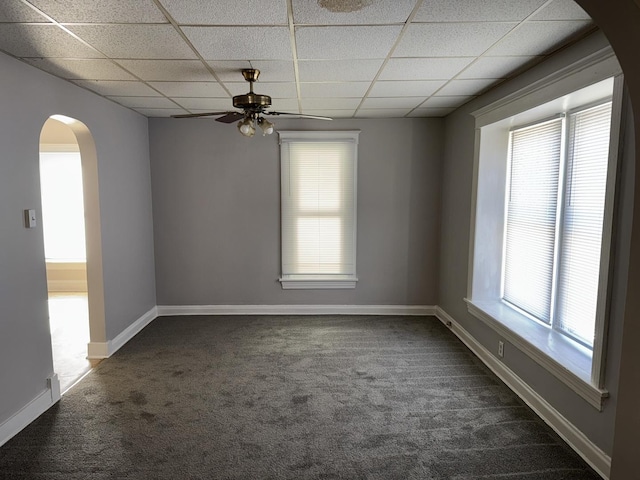 empty room with arched walkways, a paneled ceiling, a ceiling fan, baseboards, and dark carpet