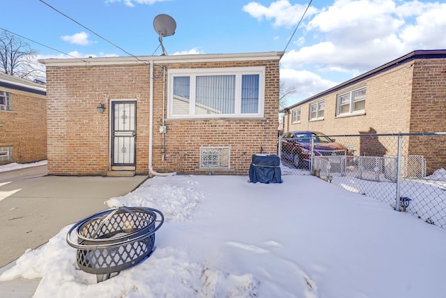 snow covered rear of property featuring brick siding, fence, and a fire pit