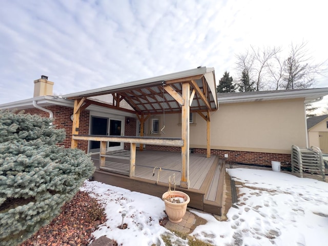 snow covered rear of property with stucco siding, a wooden deck, a chimney, and brick siding