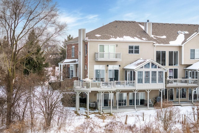 snow covered rear of property featuring a shingled roof, a sunroom, a chimney, and a deck