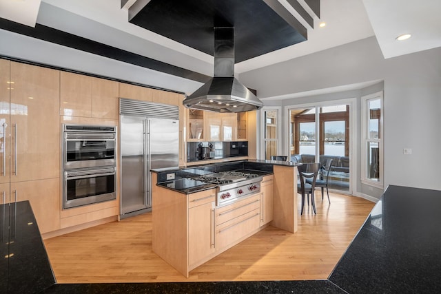 kitchen with light wood-style flooring, stainless steel appliances, light brown cabinetry, dark stone countertops, and island exhaust hood