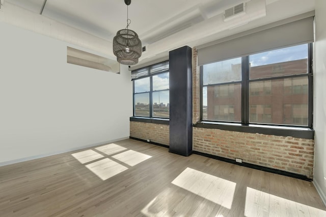 empty room with visible vents, a raised ceiling, baseboards, brick wall, and light wood-style floors