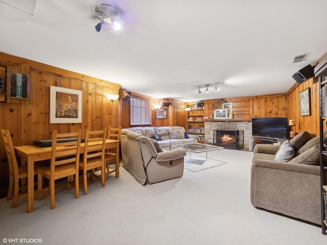 carpeted living room with a stone fireplace, wood walls, track lighting, and visible vents