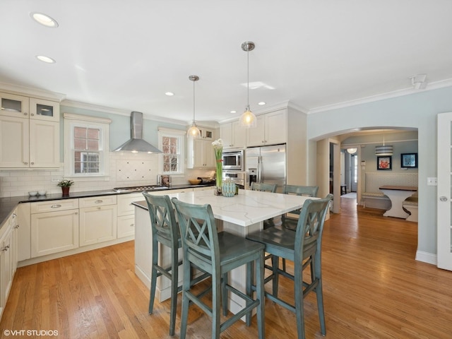 kitchen with arched walkways, ornamental molding, stainless steel appliances, wall chimney exhaust hood, and a center island