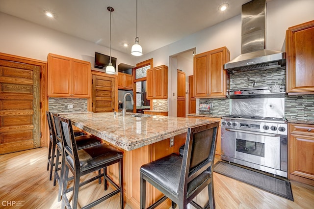 kitchen with light stone counters, a kitchen island with sink, a sink, wall chimney exhaust hood, and stainless steel range with gas stovetop