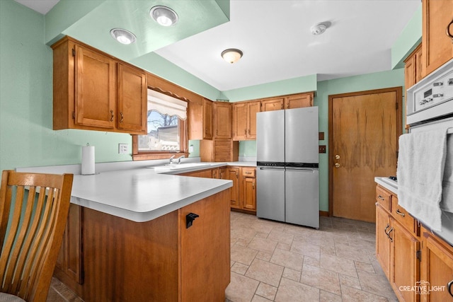 kitchen with light countertops, a peninsula, freestanding refrigerator, brown cabinetry, and a sink