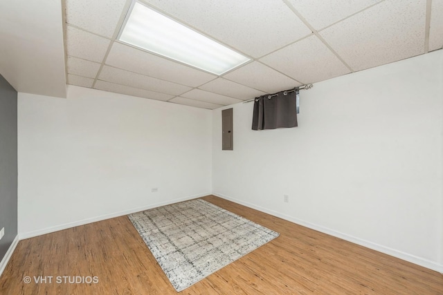 finished below grade area featuring a paneled ceiling, electric panel, baseboards, and wood finished floors