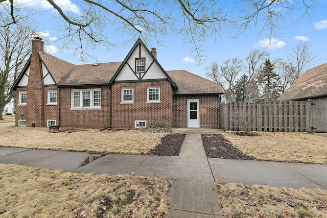 english style home featuring roof with shingles, a chimney, fence, and brick siding