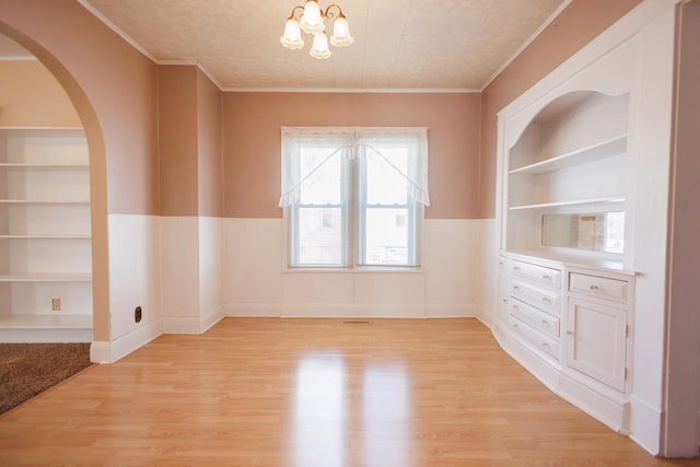 empty room with built in features, wainscoting, light wood-type flooring, an inviting chandelier, and crown molding