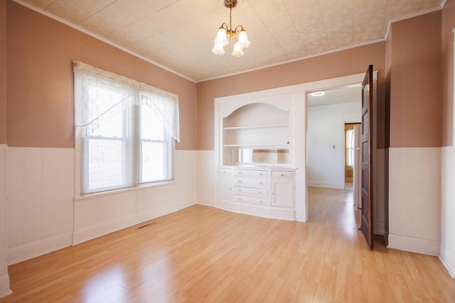 unfurnished dining area with built in features, a wainscoted wall, light wood-style flooring, an inviting chandelier, and crown molding