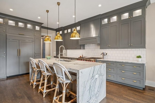 kitchen featuring light stone countertops, premium range hood, a center island with sink, and decorative backsplash