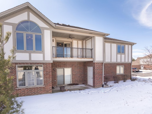 snow covered rear of property with brick siding, a balcony, and stucco siding