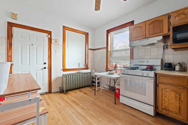 kitchen with radiator heating unit, light wood-type flooring, black microwave, under cabinet range hood, and white gas range oven